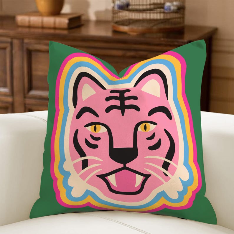 May include: A square cushion with a green background. It features a pink tiger face with black stripes and yellow eyes. The tiger face is outlined with a rainbow of colours, including pink, yellow, blue, and orange. The cushion is on a white sofa.