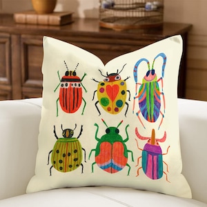 May include: Cream-colored square throw pillow featuring a colorful illustration of six whimsical beetles. The beetles are in various colors, including red, green, yellow, and blue, with detailed patterns. The pillow is on a white couch.