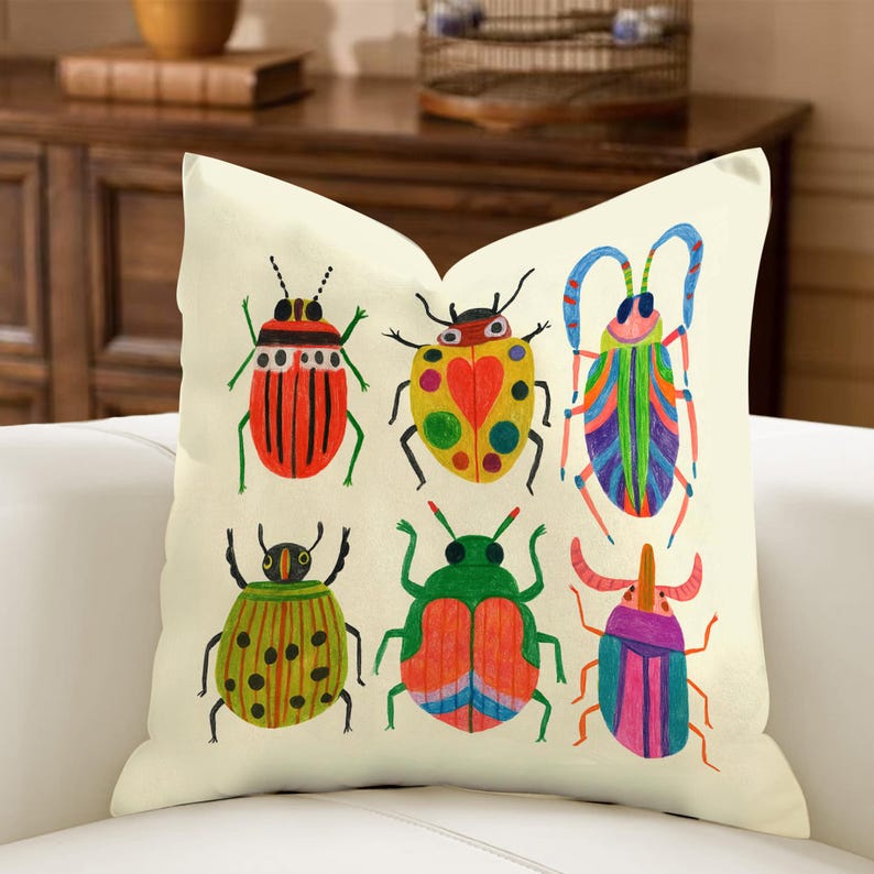 May include: Cream-colored square throw pillow featuring a colorful illustration of six whimsical beetles. The beetles are in various colors, including red, green, yellow, and blue, with detailed patterns. The pillow is on a white couch.
