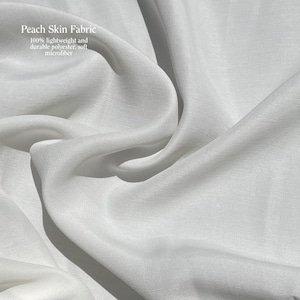Puede incluir: Primer plano de tela blanca de piel de melocot&oacute;n. La tela es 100% poli&eacute;ster ligero y duradero, microfibra suave. La tela est&aacute; drapeada en pliegues suaves, creando sombras y reflejos. El texto "Peach Skin Fabric" es visible en la esquina superior izquierda.