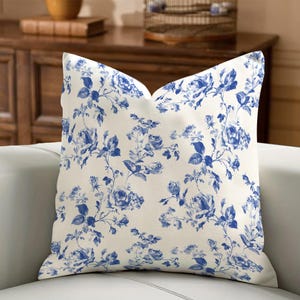 Può includere: Un cuscino quadrato con sfondo color crema e un motivo floreale blu. Il motivo presenta rose e foglie, creando un design classico ed elegante. Il cuscino è posto su un divano bianco. Le misure sono in centimetri.
