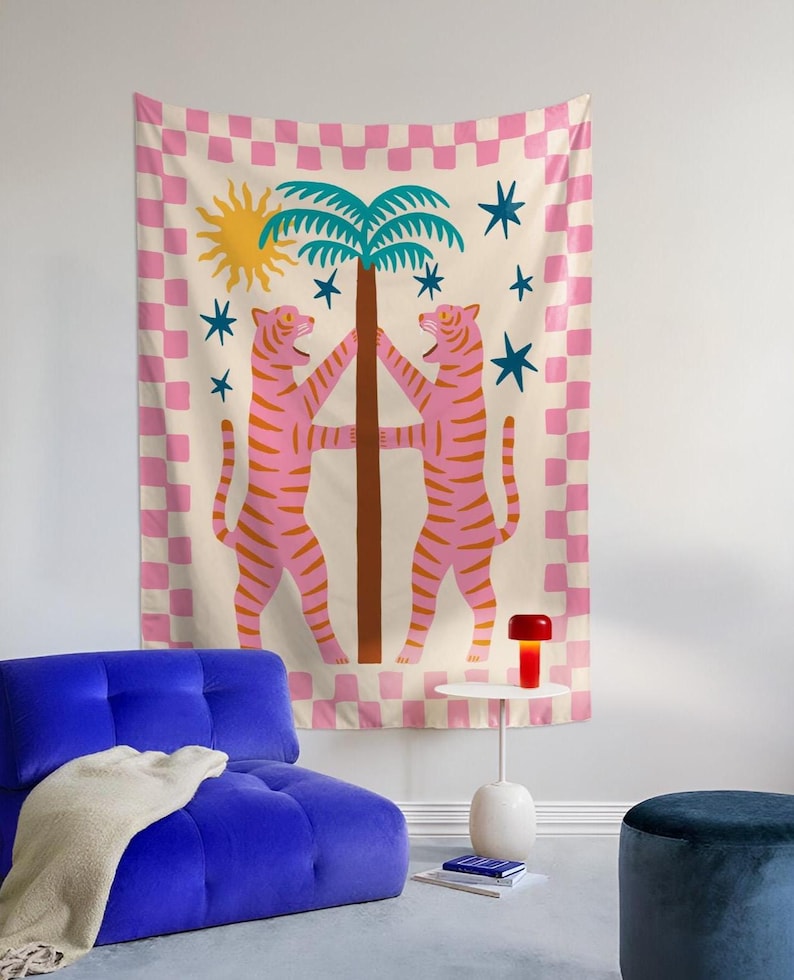 Puede incluir: Un tapiz con dos tigres rosas con rayas naranjas que flanquean una palmera marr&oacute;n. El fondo es crema con un borde a cuadros rosa y blanco. Un sol amarillo y estrellas azules completan el dise&ntilde;o. El tapiz est&aacute; colgado en una pared.