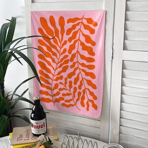 May include: A pink wall hanging with a repeating orange leaf design. The artwork is rectangular and hangs on a white shuttered door. A bottle of beer and books are visible in the foreground.