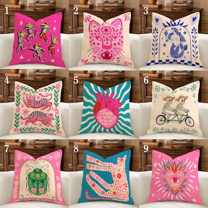 Peut inclure: Une collection de coussins décoratifs aux motifs colorés et fantaisistes. Les coussins présentent divers motifs, notamment des squelettes, un jaguar, une sirène, des chats, un cœur, un scarabée et un cœur avec du texte. La palette de couleurs comprend le rose, le bleu et la crème.