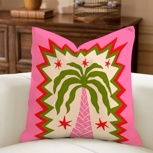 May include: A decorative throw pillow with a vibrant pink background. The pillow features a central design of a green palm tree with a pink trunk, surrounded by red stars and a zigzag border. The design is set against a cream-colored backdrop.