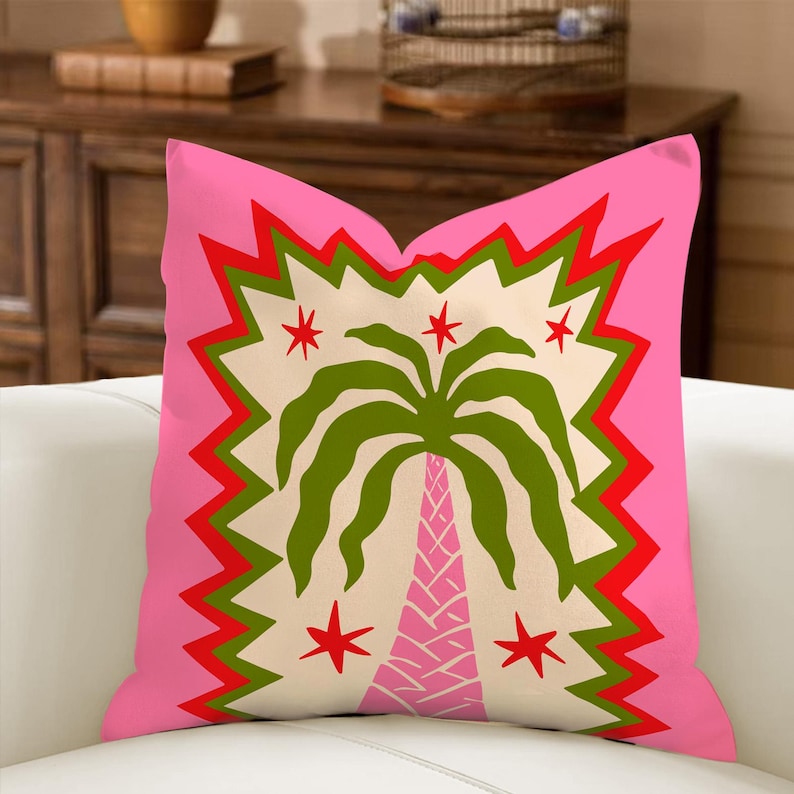 May include: A decorative throw pillow with a vibrant pink background. The pillow features a central design of a green palm tree with a pink trunk, surrounded by red stars and a zigzag border. The design is set against a cream-colored backdrop.