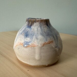 May include: A small, handcrafted ceramic vase with a rounded shape. The vase features a colour gradient, with a white base, transitioning to a light orange band, and topped with blue and brown hues. The vase sits on a wooden surface.