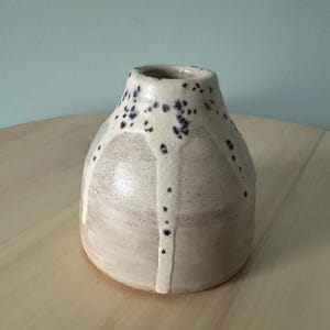 May include: A handmade ceramic vase with a rounded form and a narrow neck. The vase is mainly a light beige colour, with a white glaze that drips down the sides. Small purple dots decorate the top rim.