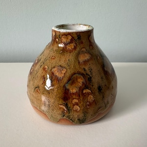 May include: A handmade ceramic vase with a rounded form and a small neck. The vase has a mottled glaze in brown, tan, and green hues, with a textured finish. The inside is white. A decorative item for the home.