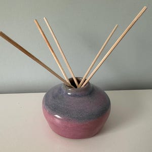 May include: A ceramic essential oil diffuser with a round, bulbous shape. The vase is a gradient of pink and grey, and contains several wooden reeds. The reeds are arranged to fan out from the top of the vase. The diffuser sits on a white surface.