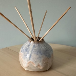 May include: A small, round ceramic vase with a light blue and beige glaze, containing several wooden diffuser sticks. The vase has a textured surface and a wavy design near the base. The sticks are arranged to fan out from the vase.