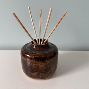 May include: A brown ceramic essential oil diffuser with several wooden reeds inserted. The diffuser has a rounded shape and a glossy finish. The reeds are light brown and arranged to fan out from the opening of the diffuser.