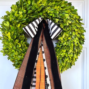 May include: A circular wreath made of dense green boxwood leaves, adorned with a large decorative bow featuring long, hanging ribbons in black, white, and orange patterns, including stripes, solid black velvet, and black polka dots on a white background.