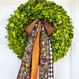 May include: A round, green wreath made of leaves, adorned with a decorative bow and streamers. The bow features orange and black striped ribbon, and the streamers have Halloween-themed patterns with skulls, pumpkins, and the word "Boo".