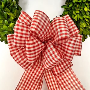 May include: A close-up of a green wreath with a large red and white gingham bow. The bow is the focal point, with multiple loops and tails. The wreath is made of small, green leaves.