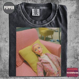 May include: Pepper-colored t-shirt with a graphic of a doll on a pink couch with a yellow pillow. The doll is wearing a pink headband and jacket. The Retroplitan logo is in the bottom right corner.