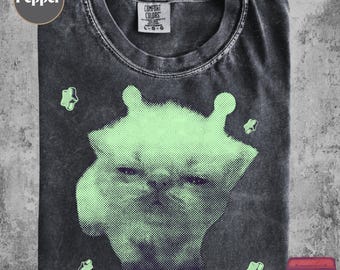 May include: A charcoal gray crew neck t-shirt with a faded, washed-out texture. The center features a large, light green halftone graphic of a cat face with two small ears, surrounded by four small, light green, four-pointed star shapes.