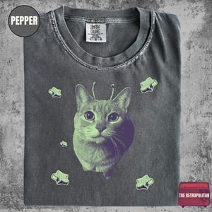 Op de afbeelding: Pepperkleurig t-shirt met een kattenafbeelding in paars en groen. De kat heeft antennes en is omringd door groene sterren. Het shirt heeft een Comfort Colors label en de tekst "THE RETROPOLITAN" is zichtbaar.
