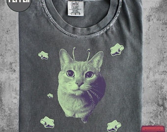 May include: Pepper-colored t-shirt featuring a cat graphic with purple and green accents. The cat has antennae and is surrounded by green stars. The shirt has a Comfort Colors tag and the text "THE RETROPOLITAN" is visible.
