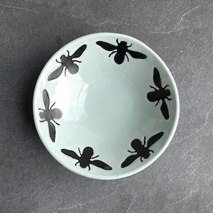 May include: A round, light mint green ceramic bowl featuring five black bee silhouettes arranged in a circular pattern around the interior rim. The bowl has a smooth, glossy finish and a simple, clean design with a wide, open basin.