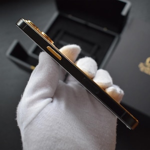 May include: A black smartphone with gold accents is held in a white gloved hand. The phone has a sleek, modern design and is encased in a protective case.