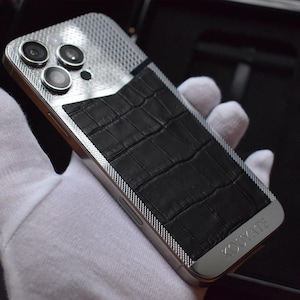 May include: A silver and black smartphone with a crocodile-embossed leather back cover. The phone has a triple camera system and the text "Caviar" is visible on the back.