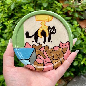 May include: A round ceramic trinket dish with a light green rim. The center features a hand-painted illustration of a yellow claw machine crane lifting a black cat above a pile of various brown and pink stuffed animal toys in a blue bin.