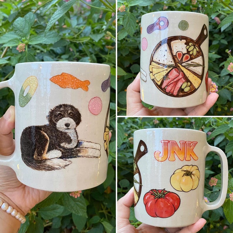 May include: Cream-colored ceramic mugs with various illustrations. One mug features a black and brown puppy, another a charcuterie board, and the third has the word "JNK" with a tomato and squash. Each mug has a large handle.