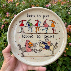 May include: A round ceramic plate with a speckled cream glaze featuring hand-painted illustrations. The top section shows five jesters holding hands with the text "born to jest". The bottom section shows two knights jousting with the text "forced: to joust".