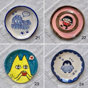 K&ouml;nnte beinhalten: Vier kleine, handbemalte Keramikplatten mit einzigartigen Designs. Eine zeigt einen Hund mit einer Sprechblase, eine andere eine Cartoonfigur, eine dritte eine gelbe Katze mit einer Tomate und die letzte einen Pinguin. Jede Platte hat einen unterschiedlich farbigen Rand.