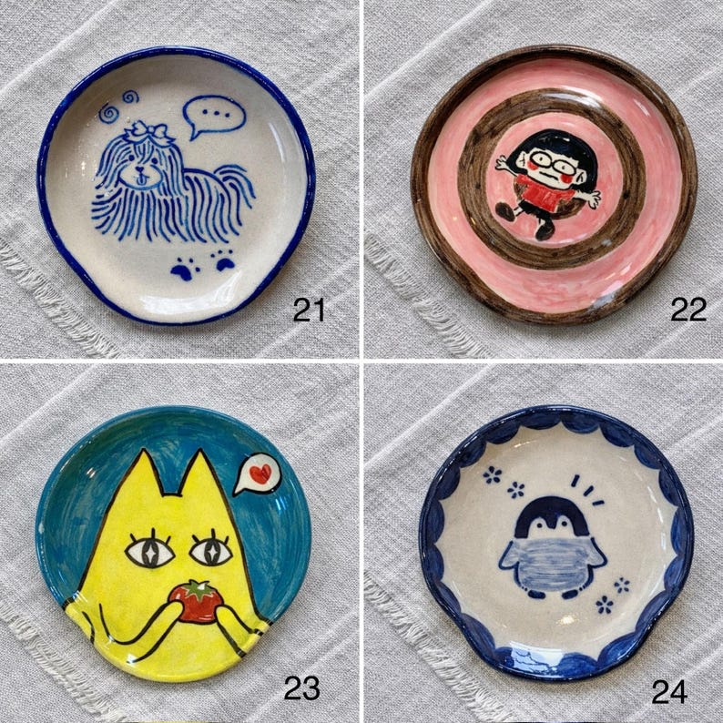 K&ouml;nnte beinhalten: Vier kleine, handbemalte Keramikplatten mit einzigartigen Designs. Eine zeigt einen Hund mit einer Sprechblase, eine andere eine Cartoonfigur, eine dritte eine gelbe Katze mit einer Tomate und die letzte einen Pinguin. Jede Platte hat einen unterschiedlich farbigen Rand.