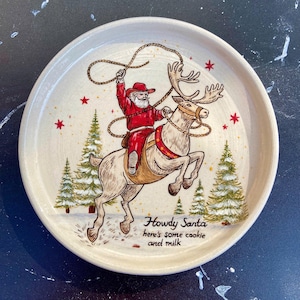 May include: A round, off-white ceramic plate featuring a hand-drawn illustration of Santa Claus in a red cowboy outfit riding a reindeer while holding a lasso. The design includes green pine trees, red stars, and the text: Howdy Santa here's some cookie and milk.