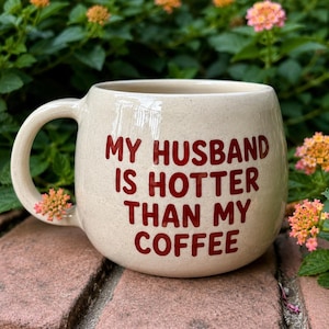 Puede incluir: Taza de cer&aacute;mica color crema con forma redondeada y asa grande. La taza presenta el texto "MY HUSBAND IS HOTTER THAN MY COFFEE" en letras rojas. Ideal para caf&eacute; o t&eacute;.