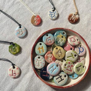 Könnte beinhalten: Handgefertigte Keramik-Anhänger-Halsketten mit einzelnen Buchstaben. Die Anhänger sind in verschiedenen Farben erhältlich, darunter Rosa, Blau, Grün und Braun, mit schwarzen Kordeln. Einige Anhänger befinden sich in einer Schüssel mit rotem Rand.