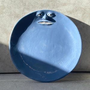 Puede incluir: Un plato de cerámica azul de forma circular que presenta un rostro humano esculpido integrado en el borde superior. El rostro tiene ojos definidos, nariz y una boca ancha con dientes blancos visibles. La superficie es de color azul mate.