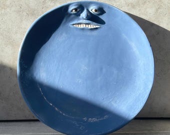 May include: A circular blue ceramic plate featuring a sculpted human face integrated into the upper rim. The face has defined eyes, a nose, and a wide mouth with visible white teeth. The surface of the plate is a solid matte blue color.