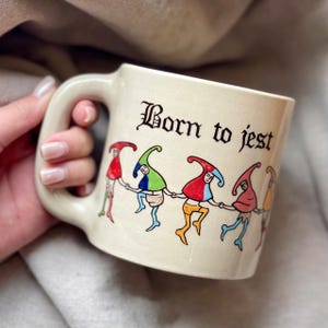May include: Cream-colored ceramic mug with a large handle. The mug features the phrase "Born to jest" in black gothic lettering. Below the text is a line of cartoon jesters in colorful outfits, holding hands.