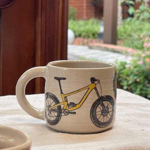 Op de afbeelding: Beige keramische mok met een illustratie van een zwarte en gele mountainbike. De mok heeft een gebogen handvat en een cilindrische vorm. Het fietsontwerp heeft zwarte banden en een geel frame.