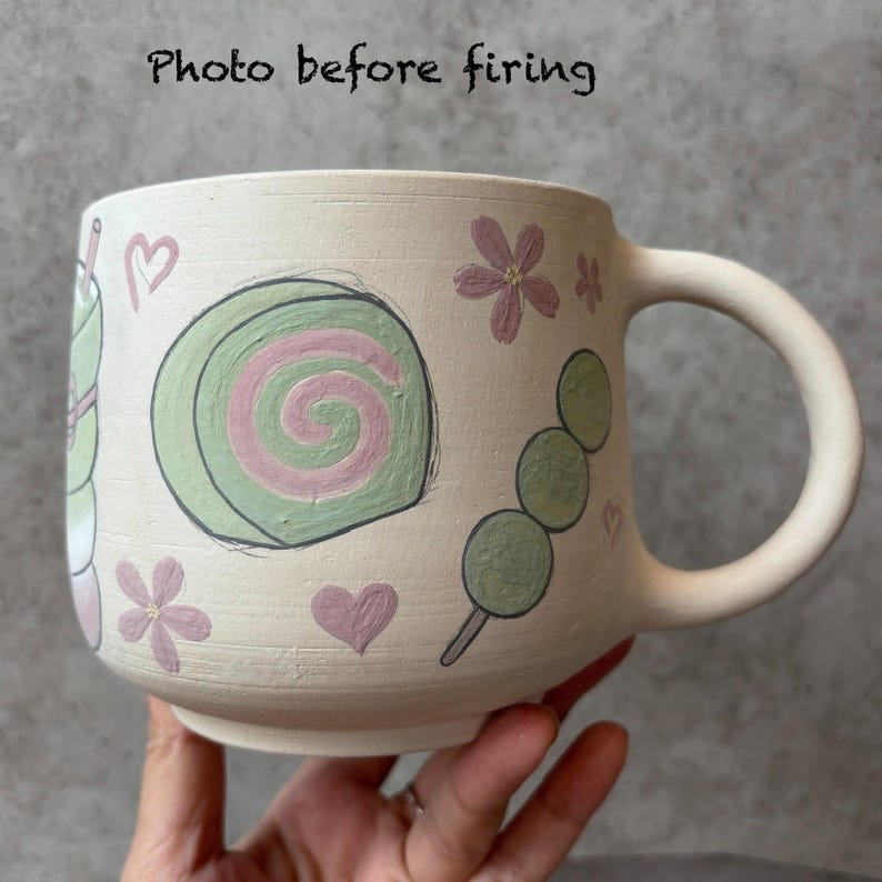 May include: A cream-colored ceramic mug with hand-painted illustrations of Japanese sweets, including a green and pink swirl cake, dango, and flowers. The mug has a large handle and the text "Photo before firing".