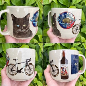 May include: Four ceramic mugs with unique designs. One mug features a black cat with different colored eyes and the text "Armadillo Den". Another mug has a bicycle and the text "Armadillo Den". The other mugs have a bicycle, a wine bottle with text, and a passport.