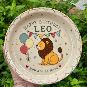 Puede incluir: Un plato redondo de cerámica con acabado crema moteado y borde festoneado. El centro presenta un león de dibujos animados, globos coloridos y banderines con el texto "HAPPY BIRTHDAY LEO" y "you are so loved" en letras oscuras.