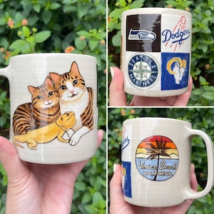 May include: Hand-painted ceramic mugs featuring various designs. One mug displays two cats and a lizard. Another mug showcases logos for the Seattle Seahawks, Los Angeles Dodgers, and Los Angeles Rams. The third mug has a Venice Beach, California design.