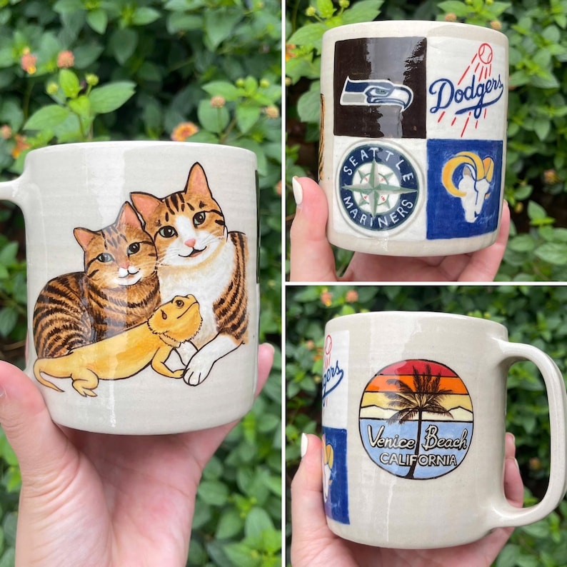 May include: Hand-painted ceramic mugs featuring various designs. One mug displays two cats and a lizard. Another mug showcases logos for the Seattle Seahawks, Los Angeles Dodgers, and Los Angeles Rams. The third mug has a Venice Beach, California design.