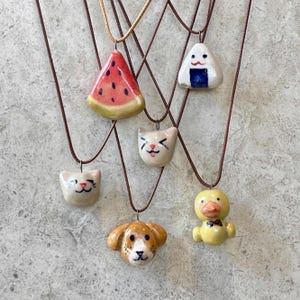 May include: A collection of ceramic pendant necklaces featuring various designs. The pendants include a watermelon slice, an onigiri rice ball, cat faces, a dog face, and a yellow duck with a bow tie. Each pendant hangs from a brown cord.