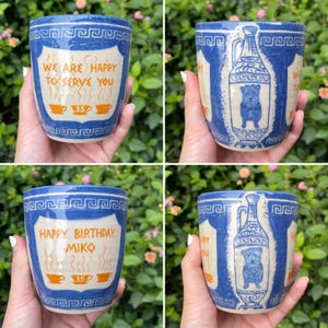 May include: A set of four ceramic mugs with a blue and white Greek key design. Two mugs feature the text "WE ARE HAPPY TO SERVE YOU" and "35" with coffee cup illustrations. The other two mugs have "HAPPY BIRTHDAY MIKO" and a dog illustration.