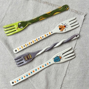 Könnte beinhalten: Vier Keramikgabeln mit handbemalten Motiven. Eine zeigt eine grüne Schlange, eine andere einen Marienkäfer, eine dritte hat einen gedrehten violetten Griff mit Sandwich-Motiv und die letzte einen blauen Käfer mit bunten Punkten am Griff.