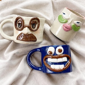 Può includere: Tre tazze in ceramica stravaganti con facce espressive. Una tazza è bianco sporco con baffi e occhiali marroni. Un'altra è blu con una grande faccia sorridente. La terza è bianco sporco con tratti del viso verdi e rosa.