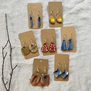 May include: A collection of handmade ceramic earrings displayed on brown cards. The earrings feature various designs, including blue bars, lemon slices, and abstract shapes in red, brown, and blue. The earrings are on silver hooks.