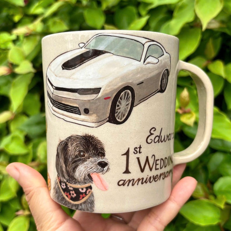 May include: A beige ceramic mug featuring a gray sports car and a dog with its tongue out. The mug is decorated with the text "Edward 1st Wedding anniversary". The mug has a curved handle.