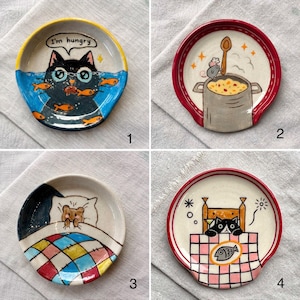 K&ouml;nnte beinhalten: Set aus vier Keramik-L&ouml;ffelablagen mit skurrilen Katzen- und Maus-Designs. Jede Ablage zeigt eine einzigartige Illustration: eine Katze mit Fischen, eine kochende Maus, eine Maus im Bett und eine Katze an einem Tisch mit einem Fisch. Die Ablagen haben farbenfrohe R&auml;nder und verspielte Bilder.